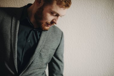 Çekici kırmızı sakallı erkek n şık takım elbise gülümseyen ve alçı duvarının yakınında dururken aşağı bakıyor