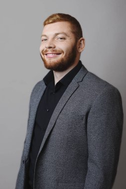 Yakışıklı kırmızı sakallı adam zarif yan görünüm uygun neşeyle gülümseyen ve gri arka plan üzerinde dururken kamera bakıyor