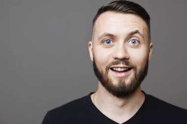 Siyah T-shirt gri arka plan üzerinde dururken heyecanlı yüz ifadesi ile kameraya bakarak yakışıklı genç adam