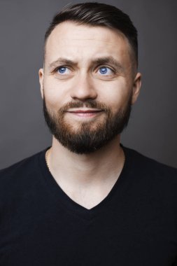 Çekici sakallı erkek sırıtışları ve gri arka plan üzerinde duran ve düşünme sırasında siyah T-shirt '