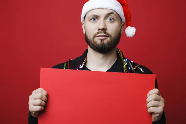 Handsome guy in Christmas hat raising eyebrow and looking at camera while standing on red background and demonstrating blank banner