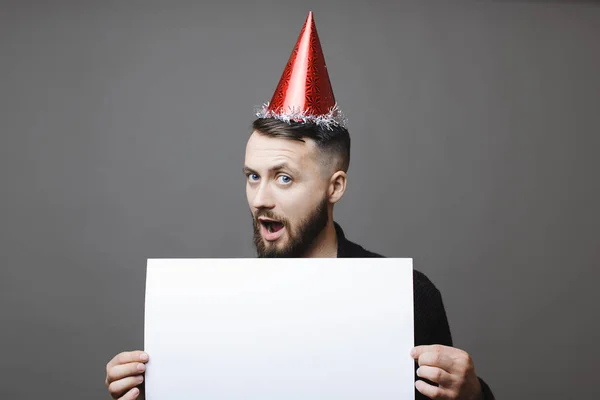 Attractive guy in red party hat holding blank banner and looking at camera with amazed face expression while standing on gray background
