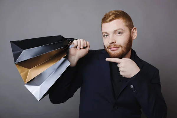 Handsome guy with ginger hair smiling and looking at camera while standing on gray background and pointing at bunch of paper bags