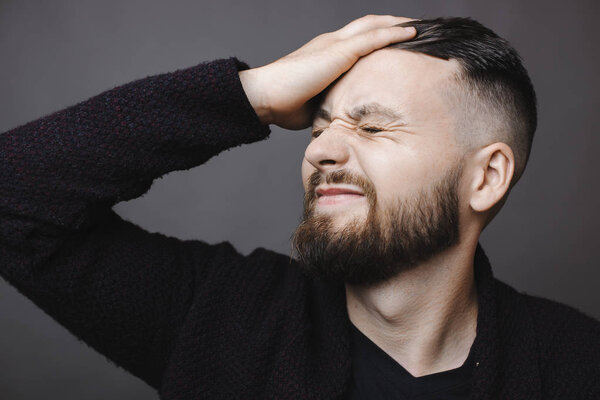 Handsome bearded guy grimacing and slapping forehead with hand while standing on gray background