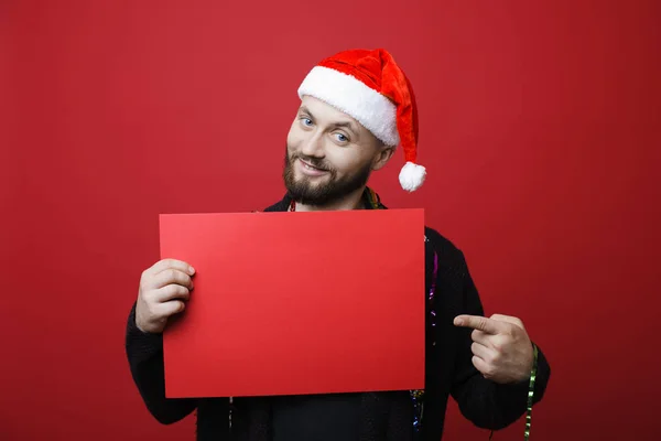 Attractive guy in Christmas hat smiling and looking at camera while standing on red background and pointing at blank banner