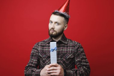 Kameraya bakarak ve küçük Doğum günü hediyesi duran parlak kırmızı zemin üzerine tutarak parti şapkası çekici genç adam