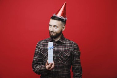 Yakışıklı genç erkek gülümseyen ve kırmızı arka plan üzerinde duran ve küçük bir hediye tutarak kameraya bakarak parti şapkalı