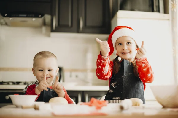 可爱的小男孩和女孩在圣诞帽微笑 并显示竖起大拇指的手势 而准备美味的糕点在舒适的厨房 — 图库照片