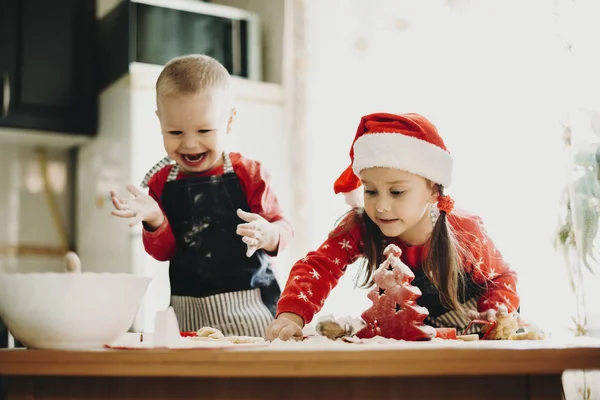 笑的婴儿男孩和女孩在圣诞老人帽子做饼干在凌乱的桌子在厨房 — 图库照片