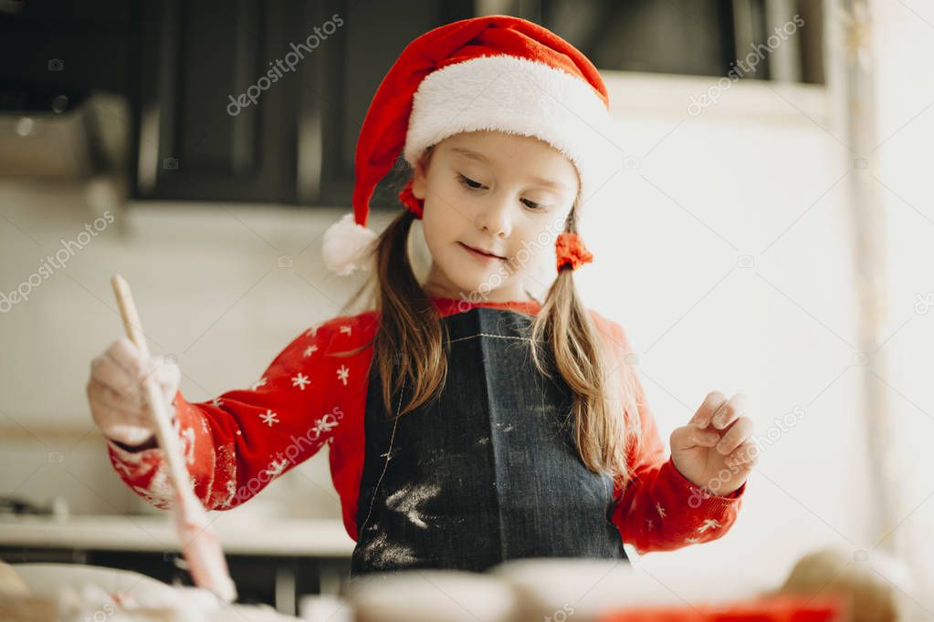 From below shot of lovely girl in apron and Christmas hat holding brush and preparing pastry while standing near kitchen table