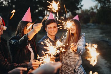 Neşeli genç arkadaş grubu yanan havai fişekleri tutarak ve kırsal kesimde inanılmaz akşam dururken gülümseyen