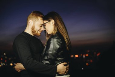 Genç adam ve kadın gözleri kapalı ve dokunaklı alınlarına gece şehir bulanık arka plan üzerinde duran ve birbirlerine sarılma tutma yan görünüm