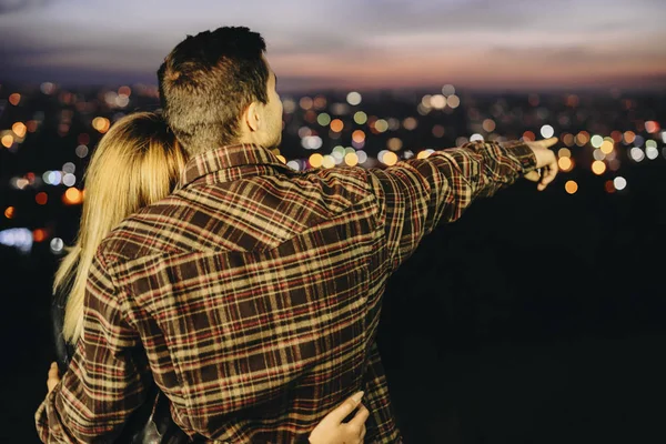 后面的看法 在格子衬衫的家伙拥抱女友和指着距离 而站在模糊的背景夜城 — 图库照片