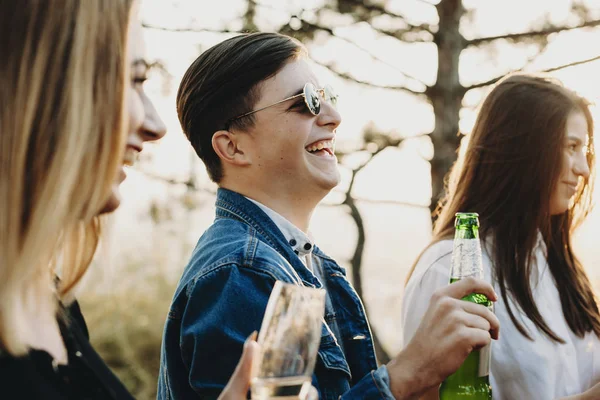 Side view of handsome young guy in stylish sunglasses holding bottle of beer and laughing while standing near women on blurred background of countryside