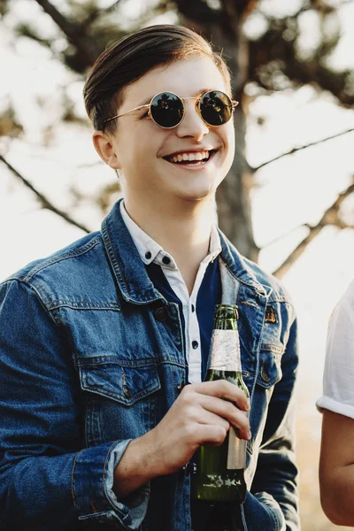 Handsome guy in casual outfit and stylish glasses drinking beer from bottle and laughing while spending time in nature on sunny day
