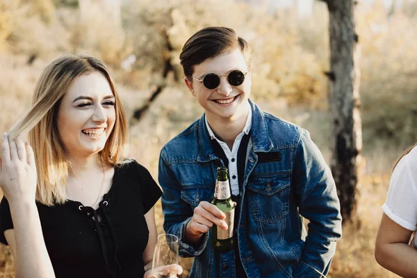 Handsome young guy with bottle of beer and lovely female with glass of champagne laughing while standing in nature on sunny day