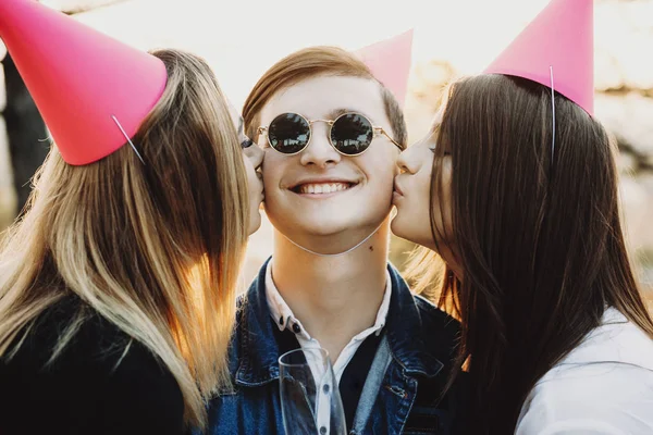 Two lovely ladies in party hats kissing cheerful guy in cheeks while having party on sunny day in nature