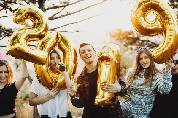 Group of young people drinking alcohol and holding balloons forming 2019 writing while standing in beautiful nature together