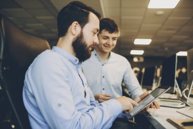 Yan görünüm yakışıklı erkek tarama modern tablet neşeli Genç meslektaşım yakınındaki office masada otururken sakallı. Sakallı adam meslektaşım yakınındaki tablet kullanma