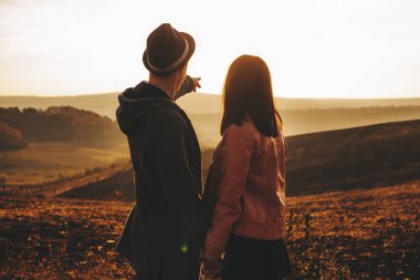 Arkadan görünüşü şık şapka muhteşem günbatımı gökyüzü anonim kız arkadaşıyla birlikte çayırda ayakta iken gösterilen genç adam. Tanınmayan adam kız için günbatımı gökyüzü işaret