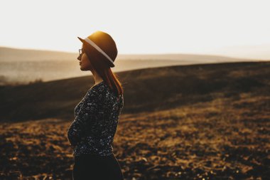 Güzel genç bayan sonbahar alan arka plan üzerinde şık şapka ayakta görkemli gün batımı sırasında yan görünüm. Sevimli kadın ayakta akşam alanındaki şapkalı