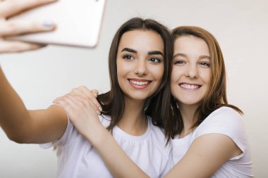 İki şaşırtıcı genç kadın gülümseyerek ve beyaz bir duvar önünde sarılma bir selfie yapıyor. Güzel kız beyaz arka plan beyaz bir akıllı telefon ile bir selfie yaparken eğleniyor.