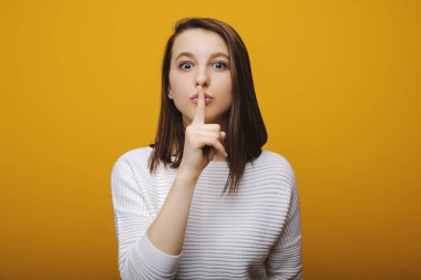 Lovely young woman dressed in casual clothes with short hair keeping a finger on her lips trying to make silence isolated on a yellow background. Shh silence sign shown by a cute female. clipart