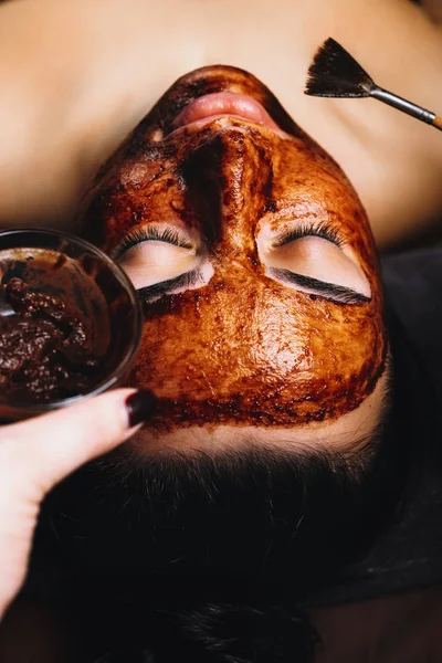 Upper view of close up young female face on which is applied chocolate mask with brush in a spa salon. Beautiful woman with closed eyes having a skin care mask with chocolate.