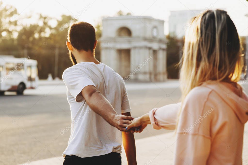 Back view of a lovely couple where man is holding her girlfriend hand and showing her where to go through the city.