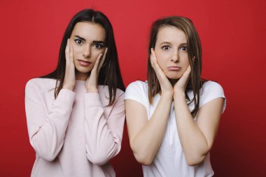 İki genç güzel kadın yanakları kırmızı bir arka plan önünde elleri ile dokunarak kamera içine şaşkın bakıyor.