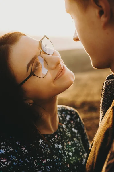 Portrait of a young cute couple where girl is looking at his boyfriend smiling wearing eyeglasses while he is looking at her against sunrise while traveling.