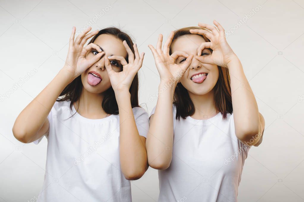 Portrait of two pretty girls having fun and showing their tongues while looking thru fingers dressed in white shirts against white wall.