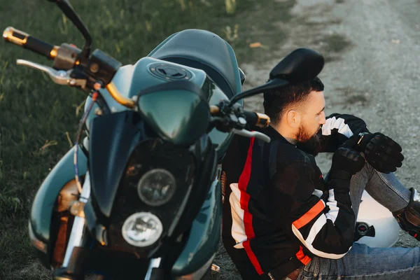 Giovane biker barbuto seduto a terra e appoggiato sulla sua moto — Foto Stock