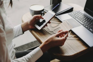 Eller internet bankacılığı veya online işlem için plastik kredi kartı ve akıllı telefon kullanarak bir pencere yakınında kahve dükkanında bir masada oturan.