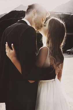 Sabah gün doğumuna karşı öpüşen güzel bir damat ve gelinin yan görünüm portresi.