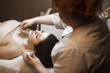 Bir sağlık spa salonunda bir kadın kozmetolojist tarafından beyaz bir cilt bakım maskesi olan genç bir güzel esmer Yan görünüm portre.