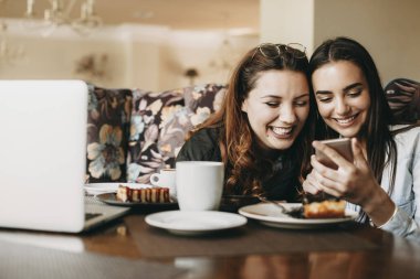 İki genç güzel beyaz kadın bir kahve dükkanında oturan bir akıllı telefon bakarken gülerek eğleniyor.