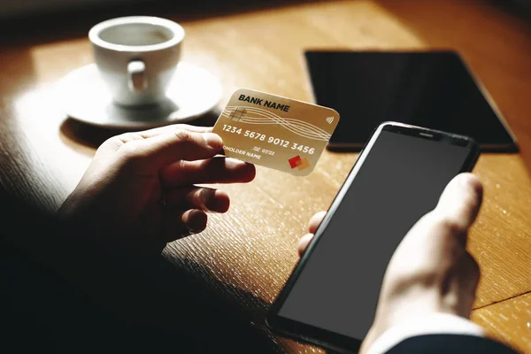 コーヒーショップでコーヒーを飲みながらオンラインバンキング用のスマートフォンとクレジットカードを使用して認識できない男性の手 — ストック写真