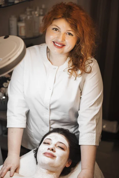 一个可爱的美容师的肖像 红头发看着相机微笑 而完成应用白色有机面具在水疗沙龙的女客户 — 图库照片