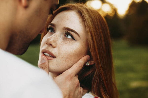 Portrait of a red haired woman with freckles and green eyes looking at her boyfriend while he is touching her lips with a finger against sunset while dating