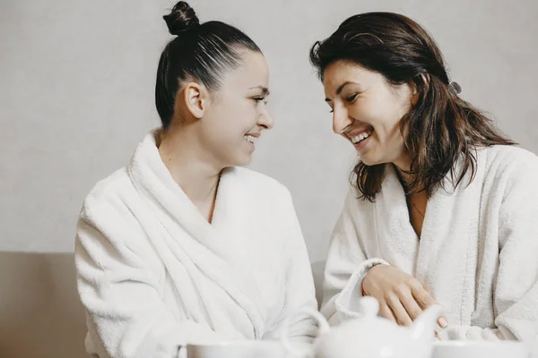 Side view portrait of two lovely young caucasian woman having fun laughing sitting in a wellness spa massage dressed in bathrobes after procedures.