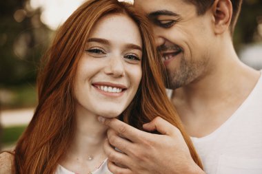 Close up portrait of a beautiful woman with freckles and red hair looking at camera laughing while her boyfriend is touching her face smiling outside. clipart