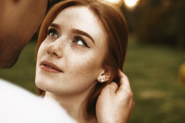 Close up portrait of a amazing red hair woman with freckles and eyes grey looking at her boyfriend smiling while he is touching her hair outside while dating. clipart
