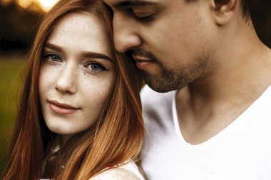 Close up portrait of a beautiful caucasian young couple embracing while she is looking at camera smiling and he is smelling her hair with closed eyes outside. clipart