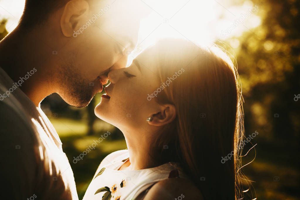 Side view portrait of a amazing caucasian couple kissing against the sunset with closed eyes white woman with freckles and red hair is smiling outside .