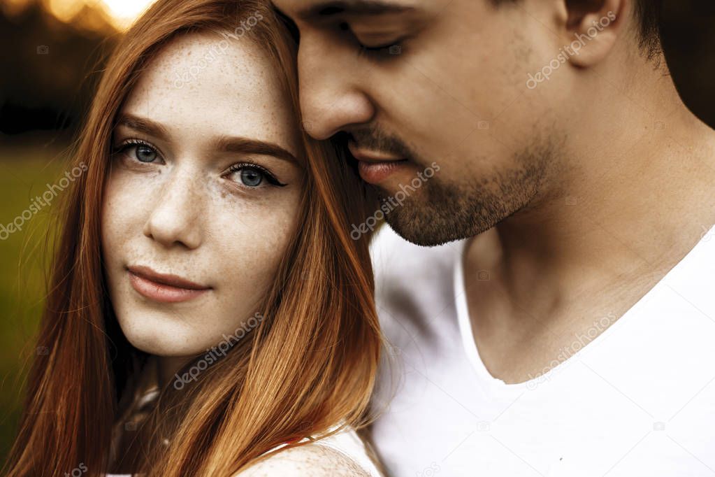Close up portrait of a beautiful caucasian young couple embracing while she is looking at camera smiling and he is smelling her hair with closed eyes outside.