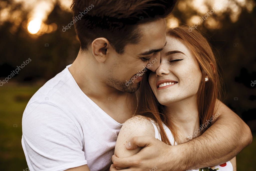 Young caucasian smiling man embracing from back his girlfriend with freckles and red hair which is with closed eyes smiling against sunset.