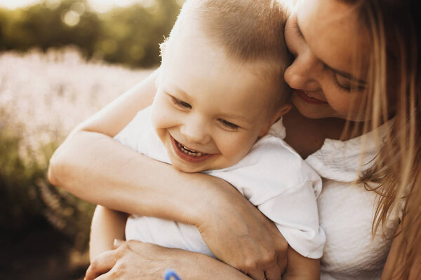 Close up shot of a cute little boy laughing while playing with his mother. Young woman embracing her son outdoor against sunset.