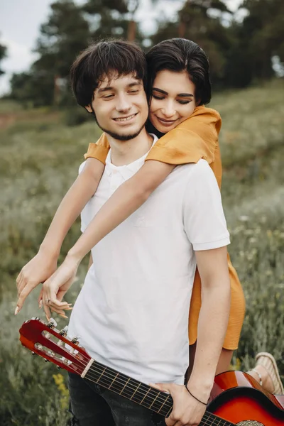 一对年轻的高加索夫妇在户外约会时 男人拿着一把吉他 女孩从背后拥抱着他的画像 — 图库照片