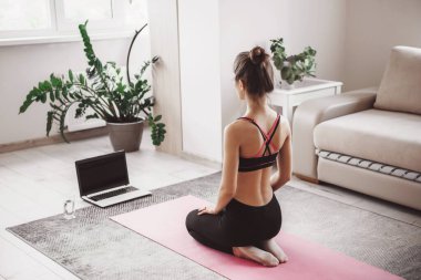 Beyaz kadın dizüstü bilgisayarın önünde oturuyor ve evde yoga yapıyor spor kıyafetleri içinde pembe bir spor halısı üzerinde.